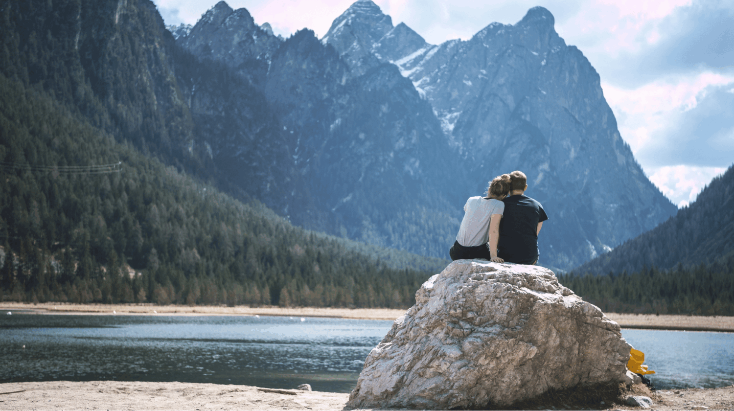 Vrouw en een man die tegen elkaar leunen zittend op een rotsblok uitkijkend over een bergmeer
